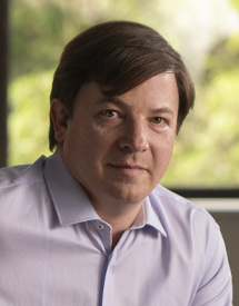 Headshot of a middle-aged white man looking at the camera with a gentle expression on his face. He's wearing a light gray/purple button up shirt sitting in front of an office window.