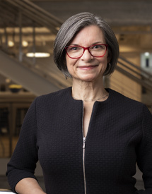 Headshot of a middle-aged white woman with glasses looking at the camera with a small smile in a black blouse standing in front of an office stairwell.