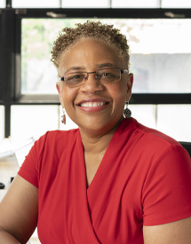Headshot of a middle-aged Black woman looking at the camera smiling and wearing a red v-neck blouse sitting in front of an office window