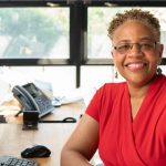 Image of a middle-aged Black woman looking at the camera smiling and wearing a red v-neck blouse sitting in front of an office window.