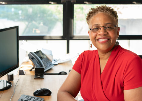 Image of a middle-aged Black woman looking at the camera smiling and wearing a red v-neck blouse sitting in front of an office window.